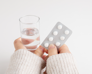 白いニットを着ている女性の手が、グラスに入った水と白い錠剤のPTPシートを持っています